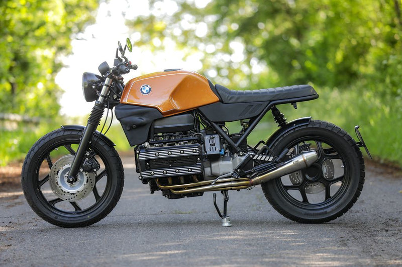 BMW K100 SE Roadster bike in vibrant orange color parked on a country road with green trees in the background.